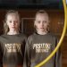 Twin girls stand in a gymnasium, each wearing a 'Positive Thoughts' shirt through a yellow hoop.